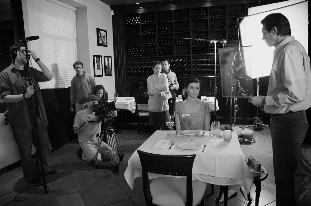 Ein Filmset mit einem Mann auf der rechten Seite, einer Frau an einem Tisch mit Tellern, Gabeln und Gläsern in der Mitte und zwei Personen, die eine Kamera bedienen, auf der linken Seite, mit Gestellen, Rahmen und einem Bildschirm im Hintergrund.