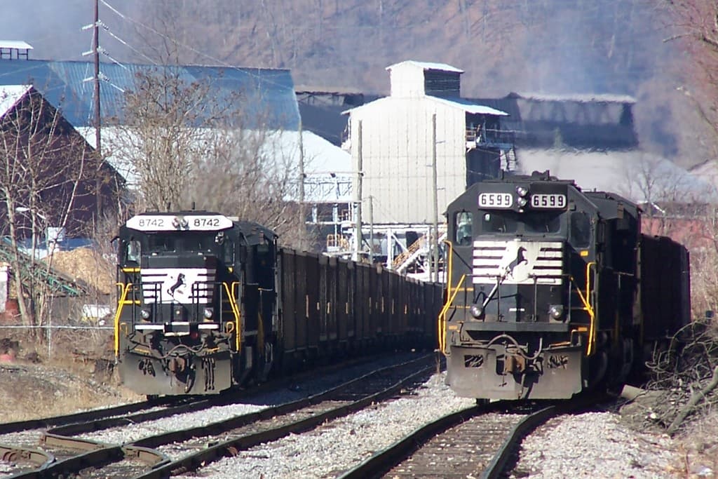 Zwei Züge auf parallelen Gleisen mit Bäumen, Strommasten und Gebäuden im Hintergrund.