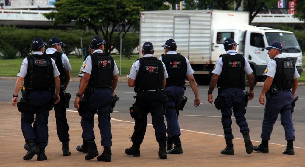 Eine Gruppe von Beamten in Uniform, einschließlich Mützen, Hemden, Schutzwesten, Uhren, Hosen und Schuhen, die auf einer Straße mit einem geparkten Lastwagen auf der rechten Seite neben einem Zaun, Gebäuden, Bäumen, Pflanzen, Polen und Bannern im Hintergrund und einer Brücke im oberen rechten Eck gehen.