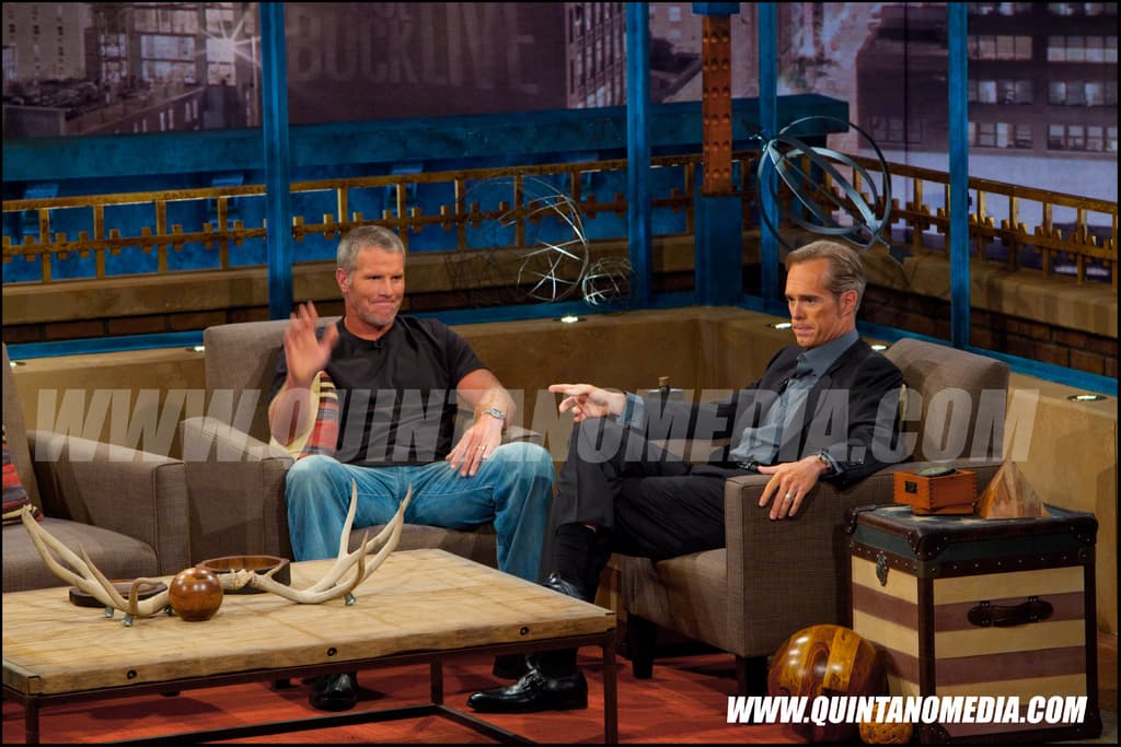 Zwei Personen sitzen auf einem Sofa in einem Medieninterview, mit einem Tisch und Spielzeugen davor, Wandplakaten dahinter und einem Mann in einem Blazer neben einem anderen Tisch mit Gegenständen und einem Holzball.