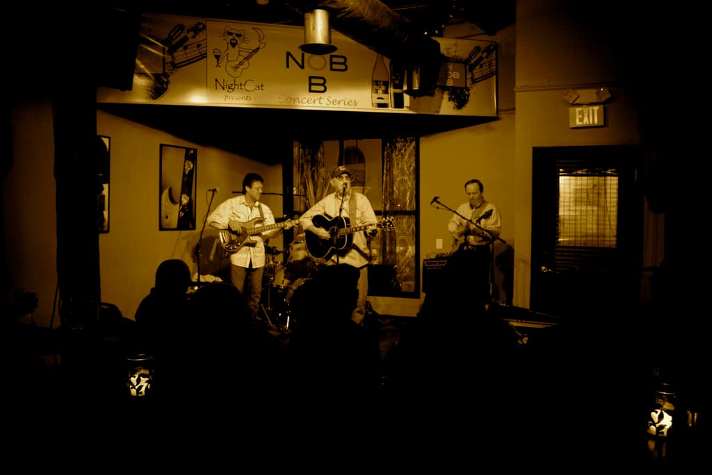 Eine Gruppe von Musikern in weißen Hemden, die Gitarren auf einer Bühne spielen, vor sich ein Mikrofon, dahinter ein Schlagzeug und über ihnen ein Banner. Die Bühne ist an eine Wand mit Gemälden gesetzt, und rechts davon ist eine Tür mit einem Schild.