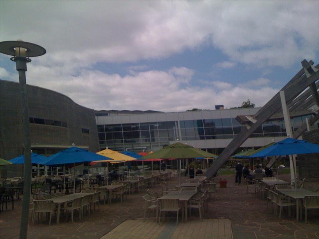 Eine Resort-Szene mit Stühlen, Tischen, Sonnenschirmen und einer Lampe im Vordergrund, Bäumen und Gebäuden in der Mitte, unter einem bewölkten Himmel.