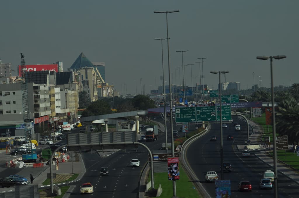 Eine Luftaufnahme einer Stadt mit Gebäuden, Türmen, Kraftfahrzeugen, Werbetafeln, Schildern, Straße, Bäumen, Straßenlaternen, Menschen, Absperrgittern und Informationsschildern unter einem klaren Himmel.