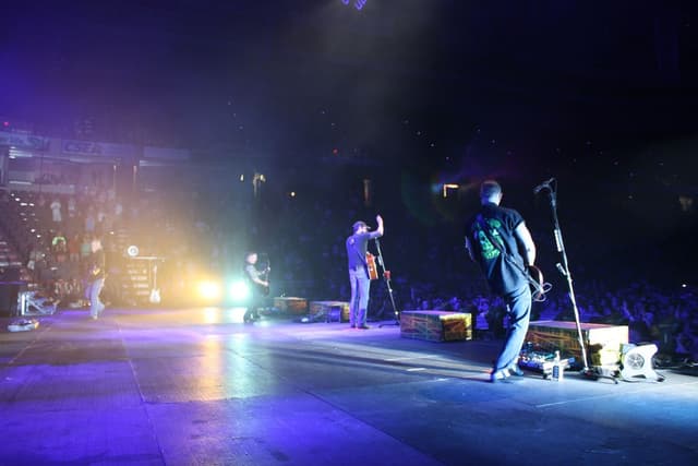 Drei Personen auf einer Bühne mit Gitarren vor einem Mikrofon, mit einer stehenden Menge im Hintergrund.