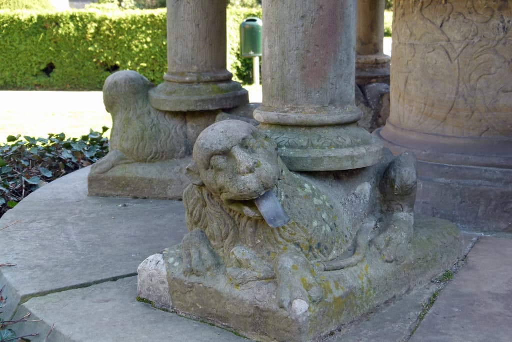 Pfähle mit Löwen-Darstellungen stehen auf einem blätterbedeckten Pfad, mit Bäumen im Hintergrund.