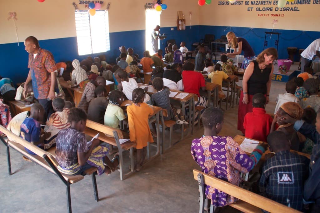 Kinder auf Bänken im Vordergrund, mit Erwachsenen, die sie führen, und einem Hintergrund mit Stühlen, einer Tür, einem Fenster, einem Tisch, Luftballons und Text oben.