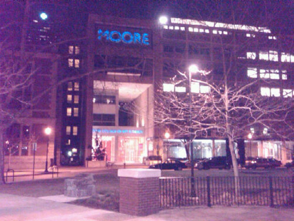 Ein Krankenhausgebäude mit mehreren Fahrzeugen, Bäumen und einem Zaun davor.