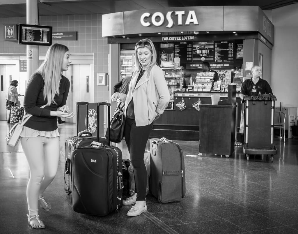 Zwei Frauen mit Gepäck vor sich, mit einem Kaffee-Shop im Hintergrund, wahrscheinlich an einem Flughafen.