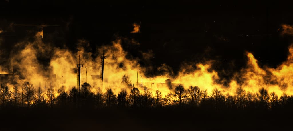 Eine dunkle Szene mit Bäumen, Strommasten, Gebäuden und Feuer.