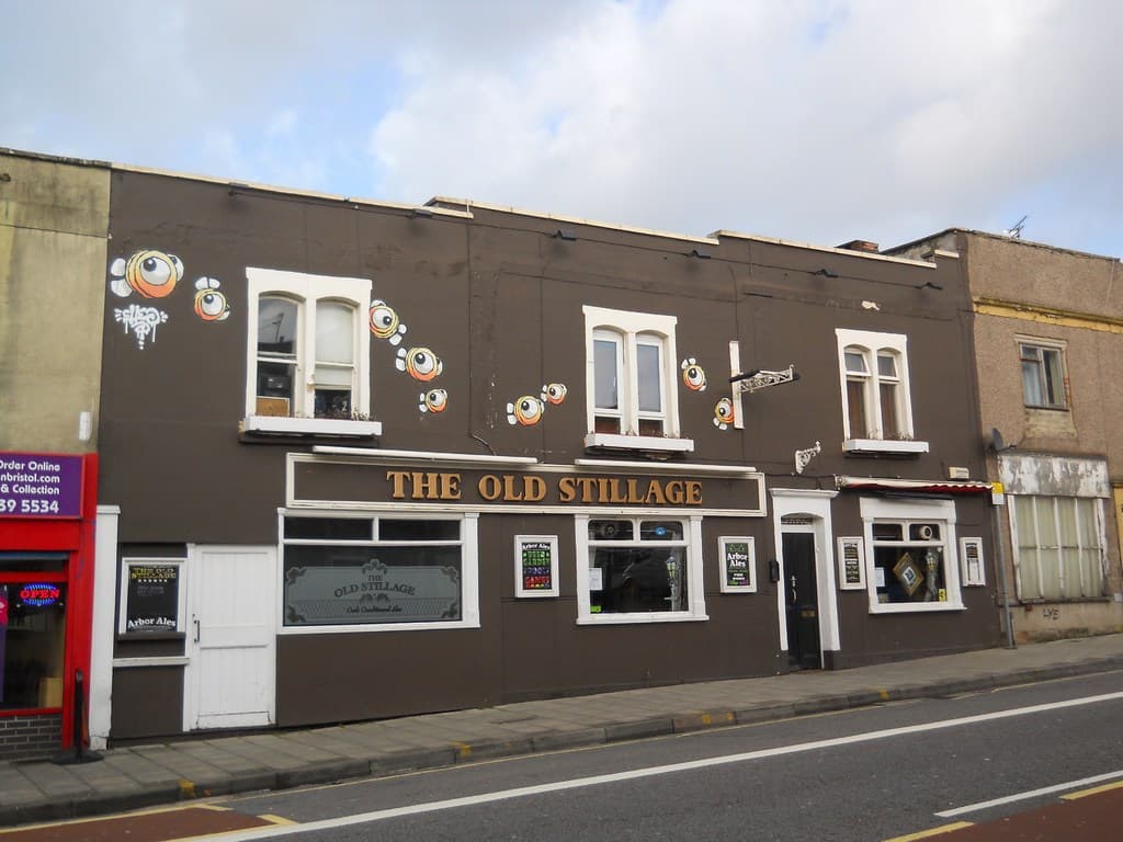 Eine Straßenansicht außerhalb der Stadt mit einer Straße, einem Gehweg und Gebäuden mit sichtbaren Türen und Fenstern, darunter eines mit der Aufschrift "Das alte nochage"; eine rote Wand und eine Glastür links, ein purpurfarbenes Schild mit Text und ein klarer Himmel darüber.