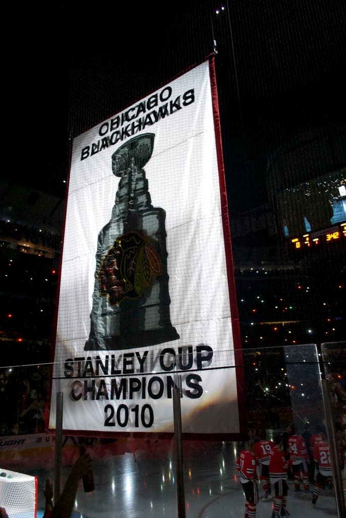 Eine nächtliche Stadionszene mit einem großen Banner in der Mitte, Zuschauern im Hintergrund, einem Glas auf dem Boden und ein paar Spielern auf dem Feld, beleuchtet von Stadionlampen.