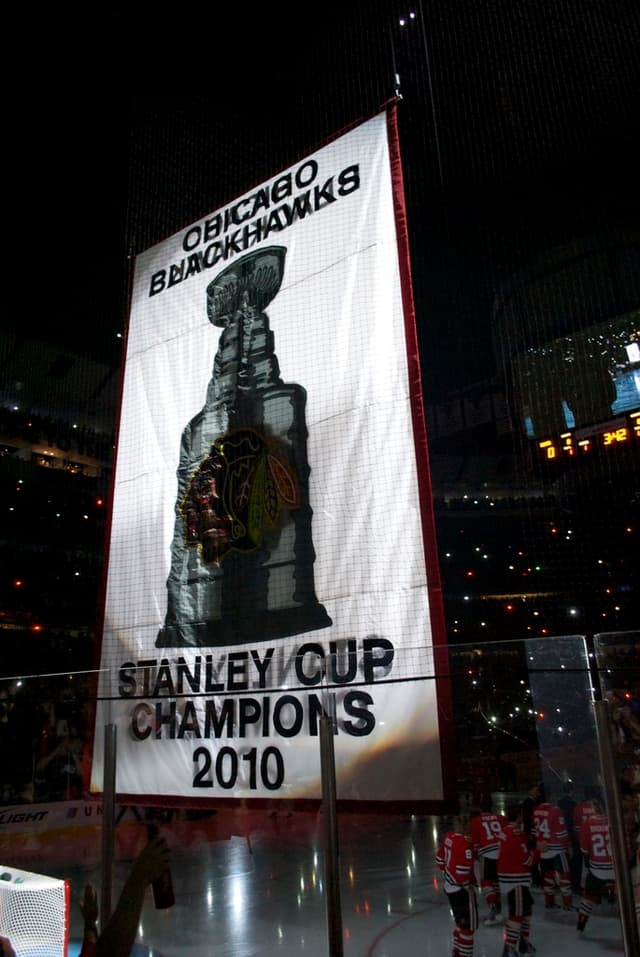 Eine nächtliche Stadionszene mit einem großen Banner in der Mitte, Zuschauern im Hintergrund, einem Glas auf dem Boden und ein paar Spielern auf dem Feld, beleuchtet von Stadionlampen.