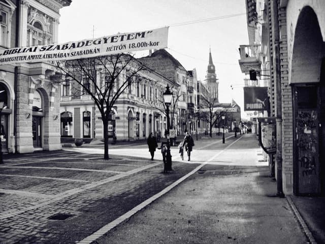 Straßenszene mit Gebäuden auf beiden Seiten, Banner oben links, Laubbäume, dekorative Laternen und einige Fußgänger