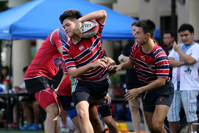 Eine Gruppe von Menschen, die Outdoor ein Spiel spielen, mit einem Jungen in der Mitte, der einen Ball hÃ¤lt, wÃ¤hrend andere stehen und im Hintergrund zuschauen, nÃ¤he eines blauen Zeltes.