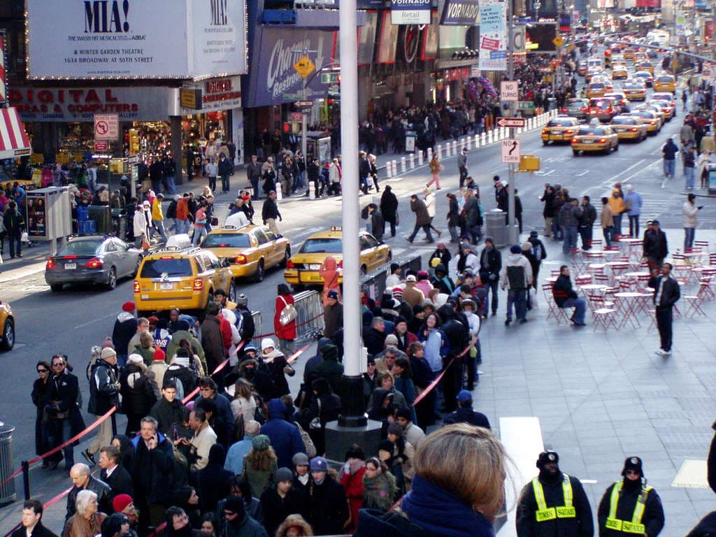 Fahrzeuge passieren eine Straße mit Fußgängern auf beiden Seiten, in der Nähe von Stühlen, Tischen, Laternen, Schildern und Ampeln, flankiert von Gebäuden mit Geschäften und Namensschildern darüber.