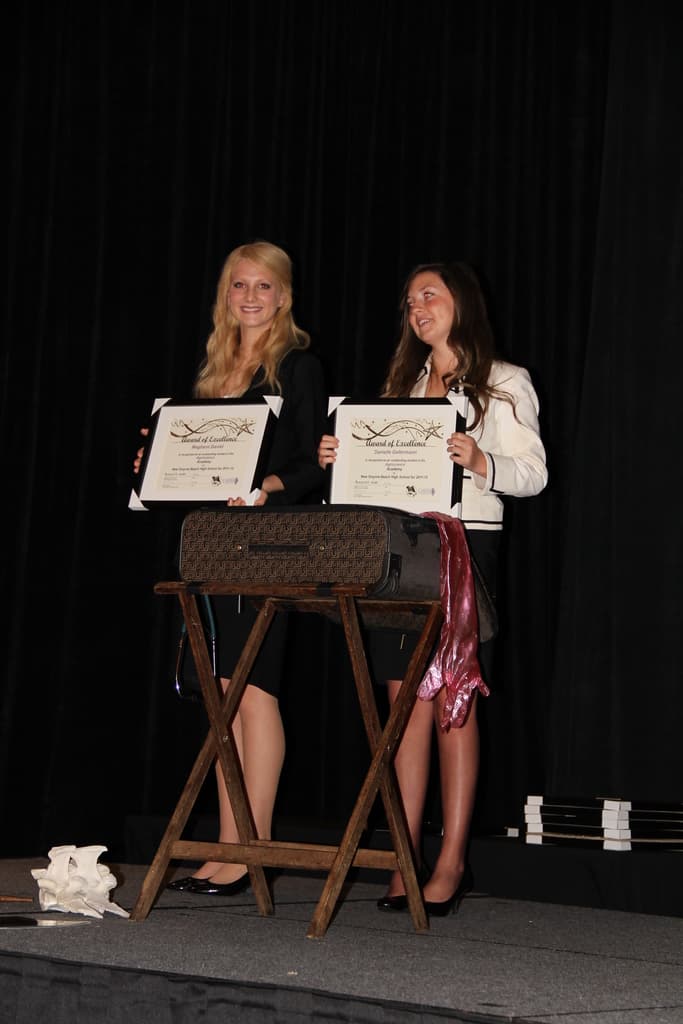Zwei Frauen, die strahlen und Zertifikate halten, vor einem Tisch mit einer Box darauf, vor einem schwarzen Vorhang.