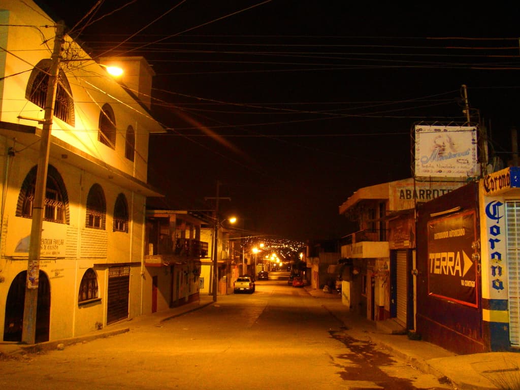 Eine Straßenszene mit Gebäuden und Strommasten auf beiden Seiten, vor den Gebäuden geparkte Fahrzeuge und eine Straße in der Mitte.