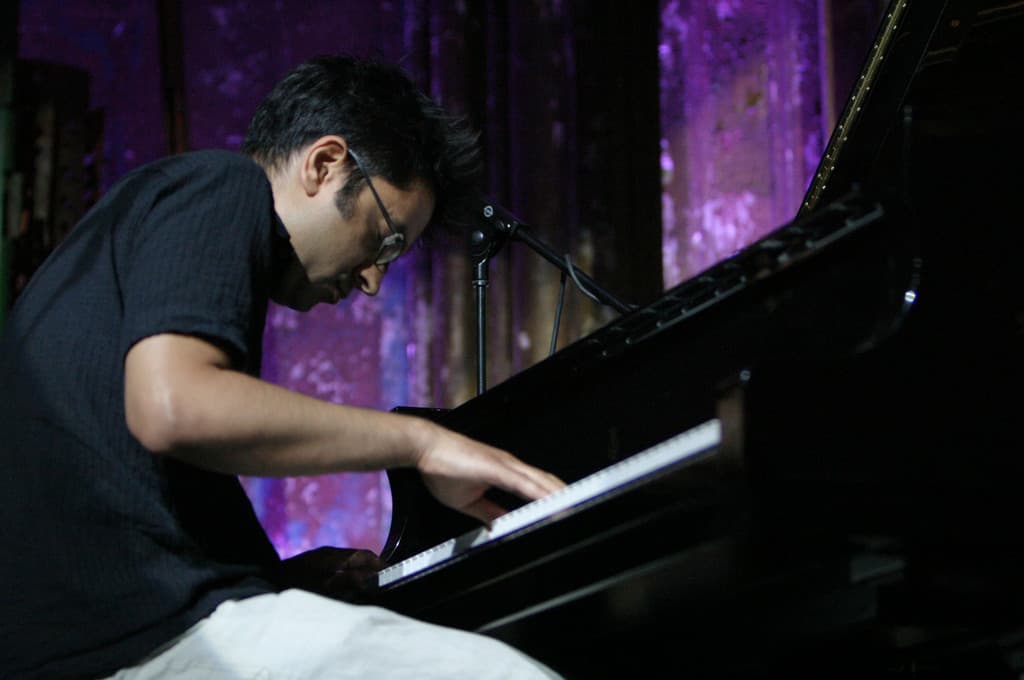 Ein Mann mit Brille spielt am Piano mit einem Mikrofonständer vor ihm und einer Wand im Hintergrund.