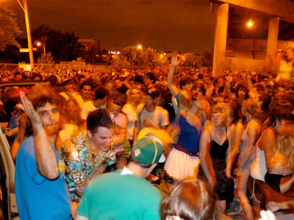 Menschen tanzen im Vordergrund bei einem Konzert mit Lichtern, Masten, Bäumen und anderen Gegenständen im Hintergrund.