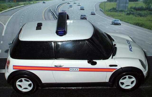 Ein weißer Polizeiwagen steht im Vordergrund mit mehreren Autos, die sich auf der Straße dahinter bewegen, umgeben von Gras und Bäumen auf beiden Seiten in einer ländlichen Umgebung.