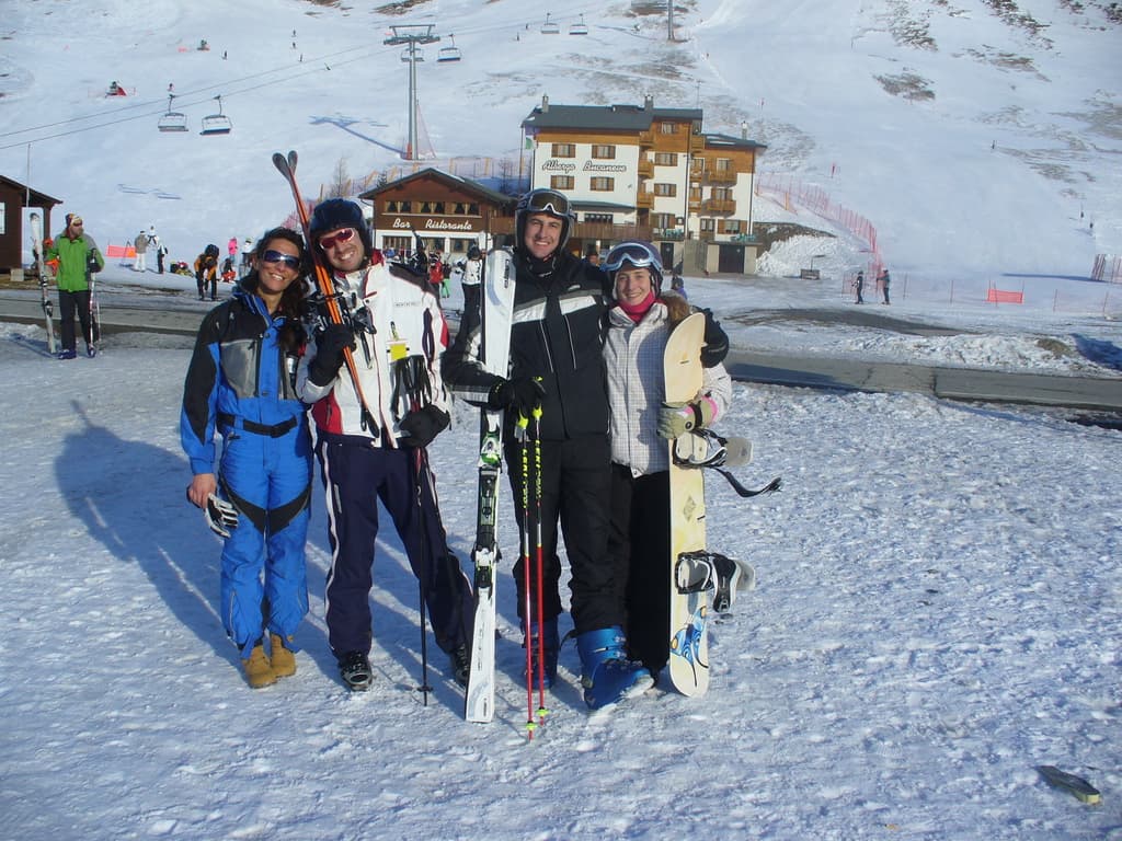 Vier Personen posieren für ein Foto mit Skaterpads und Werkzeugen, mit Gebäuden und anderen Menschen im Hintergrund, vor einer schneebedeckten Berglandschaft.