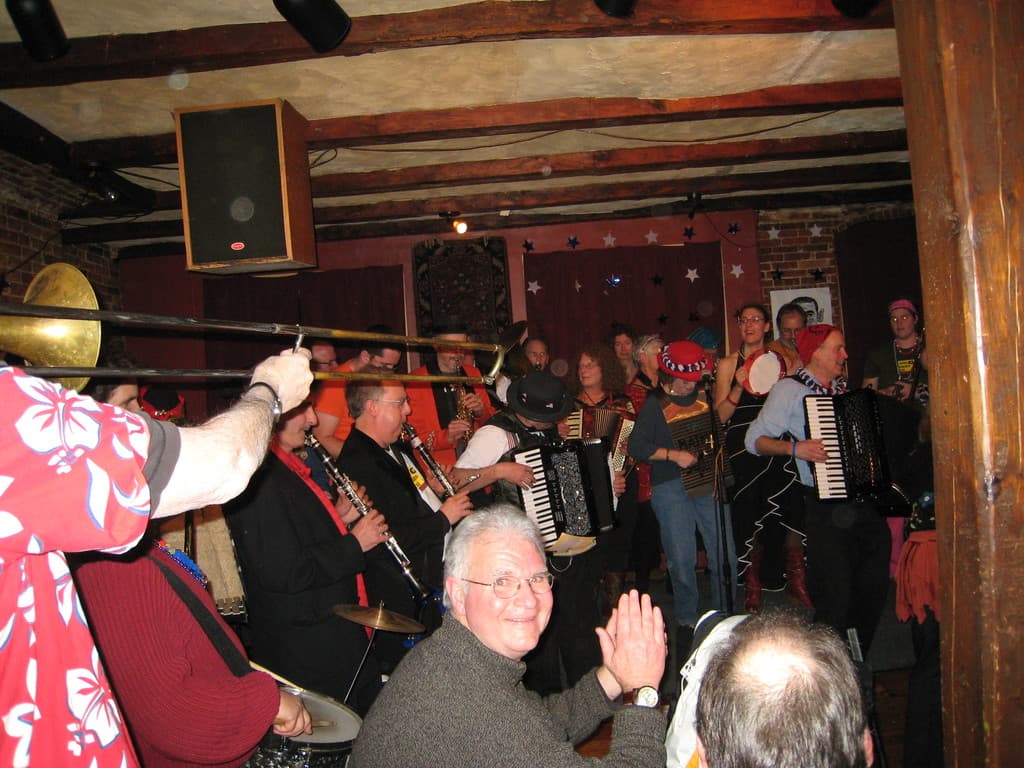 Das Innere eines Gebäudes mit sitzenden und stehenden Menschen, drei Vorhänge, ein Mikrofon mit Ständer, Musikinstrumente, Sterne, ein Lautsprecher, Kabel und eine Deckenleuchte.