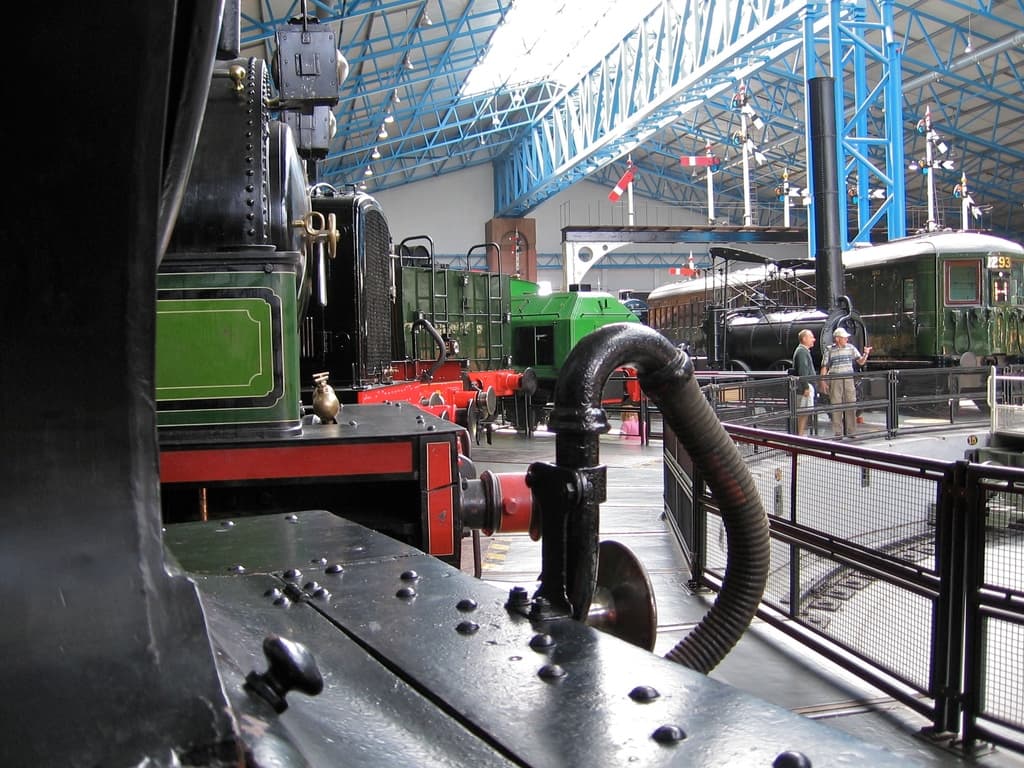 Mehrere Triebwagen stehen in einem Abstellgleis mit zahlreichen Eisenstangen oben drauf. Rechts ist ein Zaun zu sehen, neben dem ein Zug auf dem Gleis steht. Zwei Personen stehen in der Mitte neben dem Zaun.