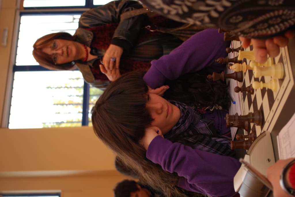 Ein Mädchen in einem lila T-Shirt sitzt beim Schachspielen, während eine Frau in einer Jacke zuschaut, mit einer Glastür und einer gelben Wand im Hintergrund.