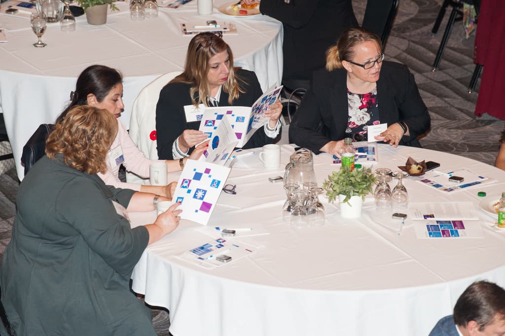 Vier Frauen auf Stühlen, eine hält ein Buch, mit einem Tisch davor, der einen Krug, Gläser, eine Tasse, einen Blumentopf, eine Pflanze, einen Teller und eine Flasche sowie mehrere andere Personen im Hintergrund zeigt.