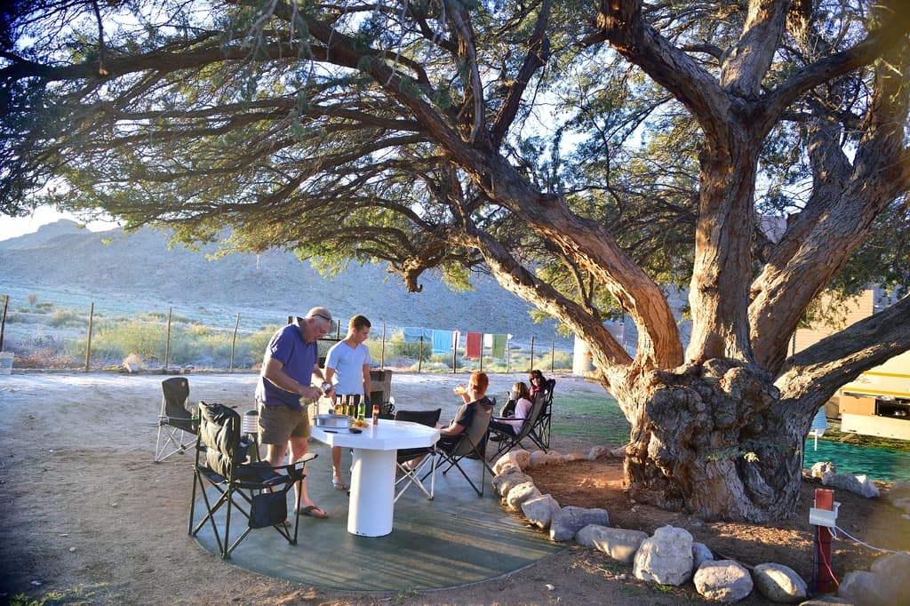 Ein Mann trägt Bierflaschen, während einige Menschen auf Stühlen sitzen, mit einem großen Baum und einem Gebäude im Hintergrund.