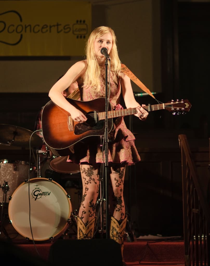 Eine Frau spielt Gitarre und singt auf der Bühne bei einem Konzert, mit einer Band und einem Bildschirm im Hintergrund und einer Tribüne unten.