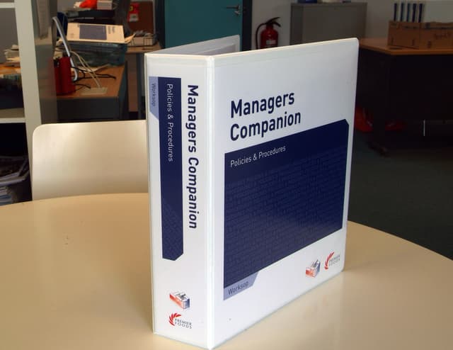 Ein Buch auf einem Tisch mit einem Stuhl dahinter, in der Nähe einer Tür, Feuerlöschern, Akten und anderen Gegenständen.