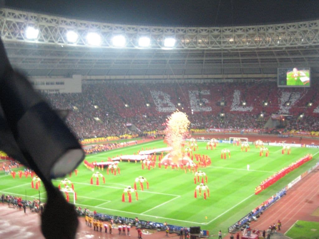 Ein Stadion mit Flutlicht, Eisengittern, Zuschauern, stehenden Personen, Werbetafeln, Anzeigetafeln, Sicherheitskameras, Fahnen und Dekorationen.