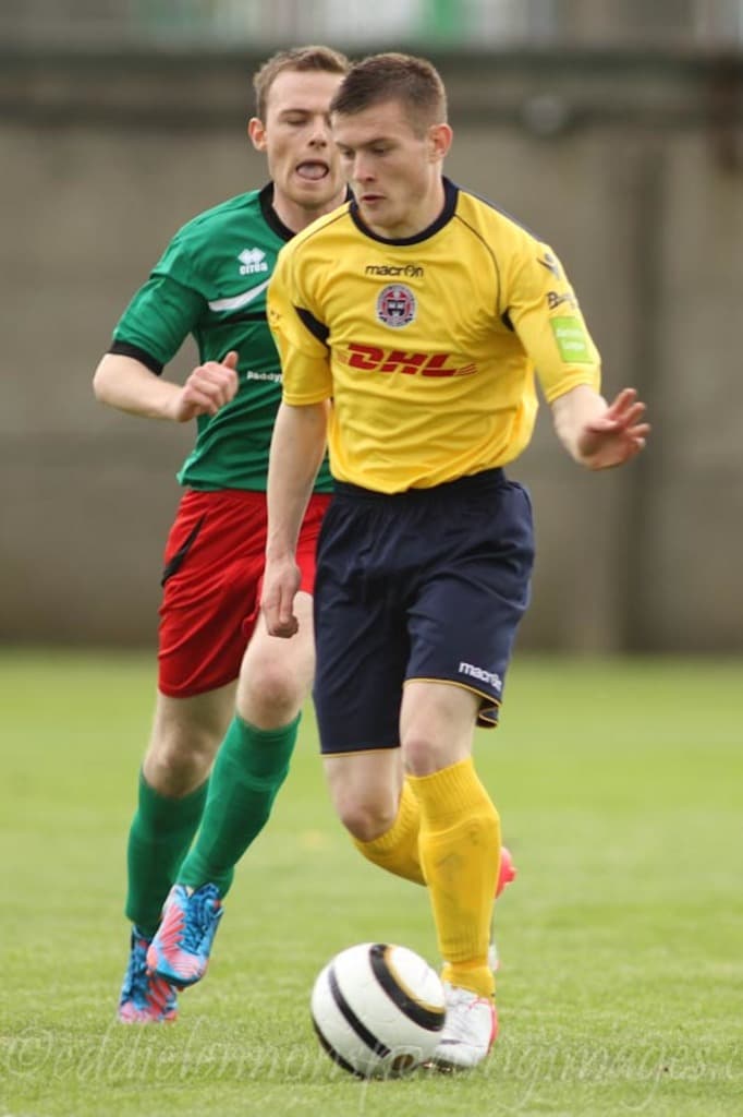 Zwei Männer bei einem Fußballspiel auf einem grünen Feld mit einem Ball, mit einem unscharfen Hintergrund und Text unten.