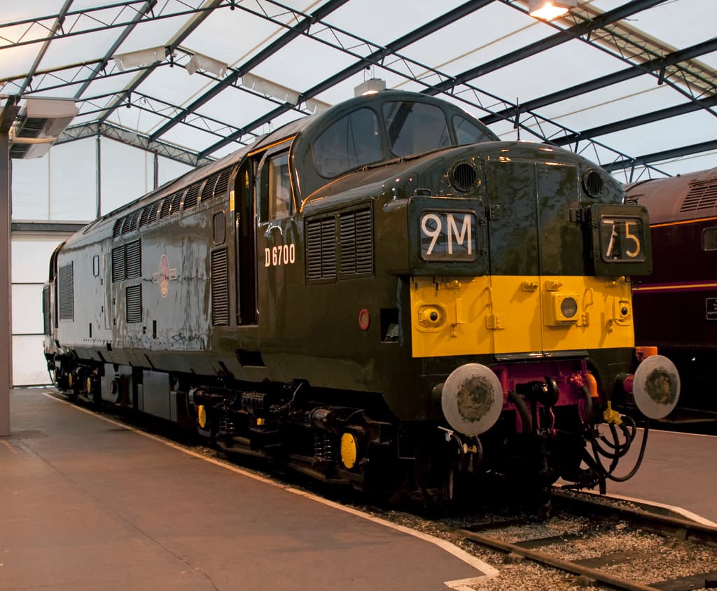 Ein Zug befindet sich auf den Schienen neben einem Bahnsteig mit einem an der Decke befestigten Mast, der Metallstangen mit Lampen trägt.