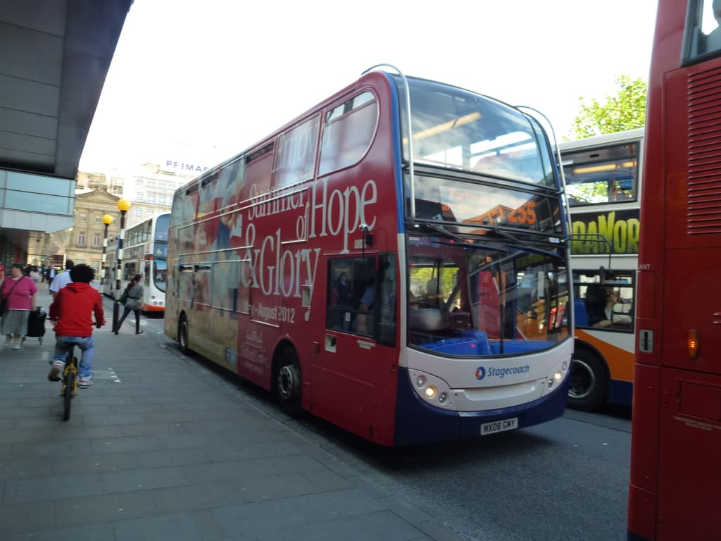 Eine Person fährt mit dem Fahrrad auf einem Gehweg mit vielen Menschen, Bussen mit Text darauf, Gebäuden und Laternen im Hintergrund.
