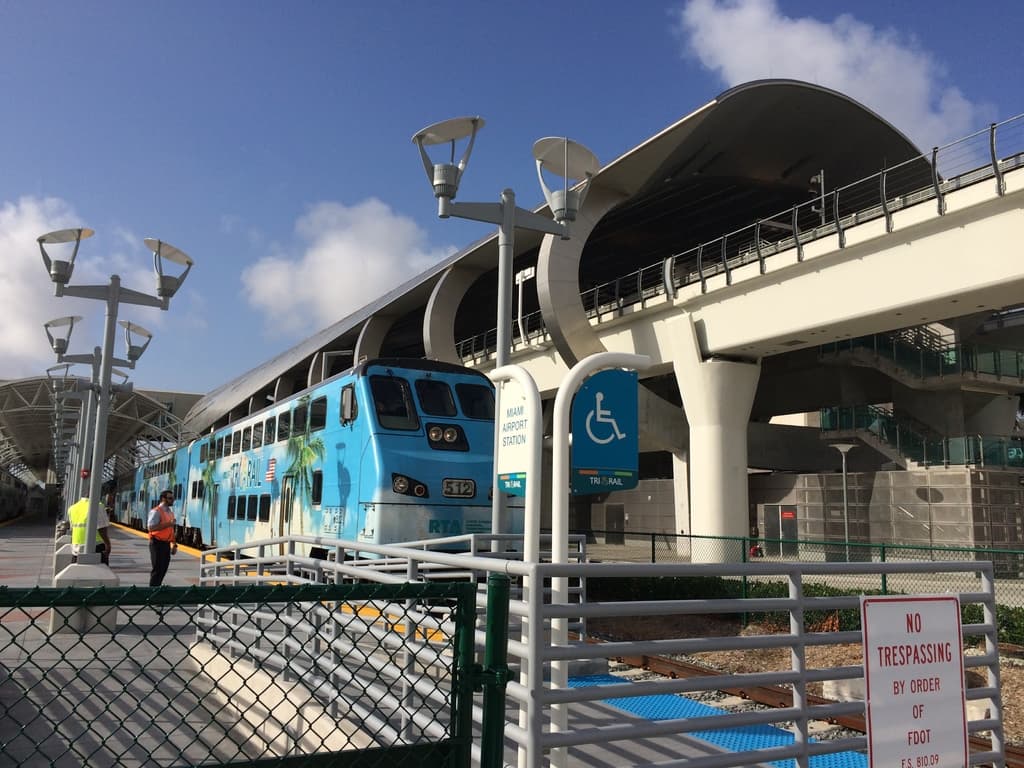 Einige Personen auf einem linken Bahnsteig mit einem Zug auf der rechten Seite, eine Brücke, ein Zaun und mehrere Pfosten.