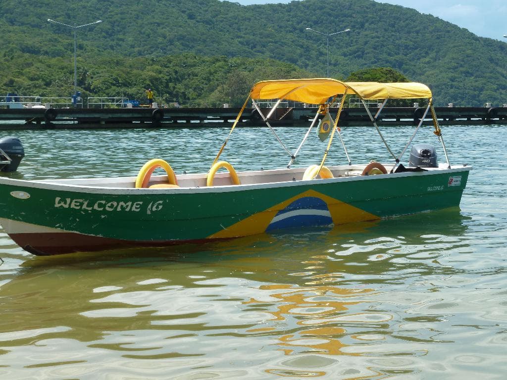 Ein Boot segelt auf ruhigem Wasser mit einer Brücke und einem baumbewachsenen Berg im Hintergrund unter einem klaren Himmel.