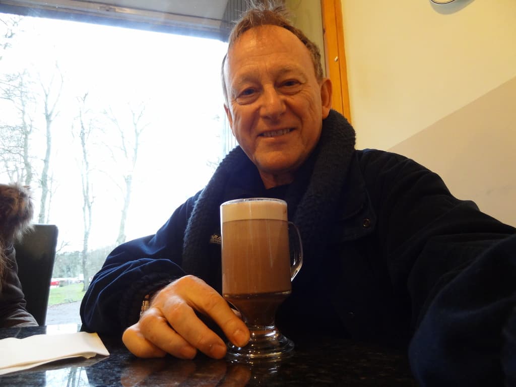 Ein Mann sitzt drinnen mit einem Bierglas vor sich, mit Bäumen und Gras sichtbar außerhalb des Raumes.