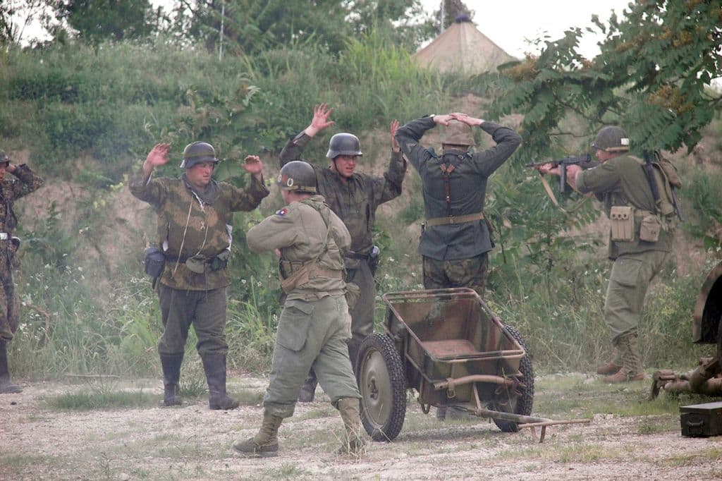 Mehrere Personen in Militäruniformen und Helmen stehen in der Nähe eines Wagens, umgeben von Pflanzen, Bäumen und Gras unter einem klaren Himmel.