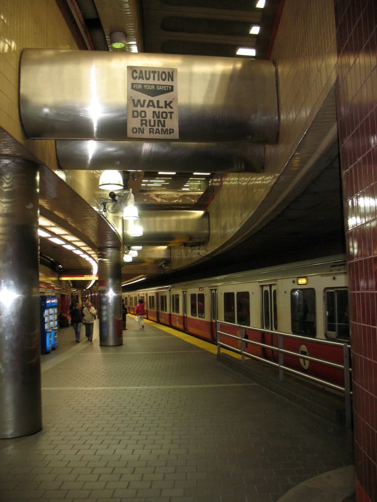 An interior view of a railway station with a train on the left and people walking on the platform, with notices visible at the top of the image.