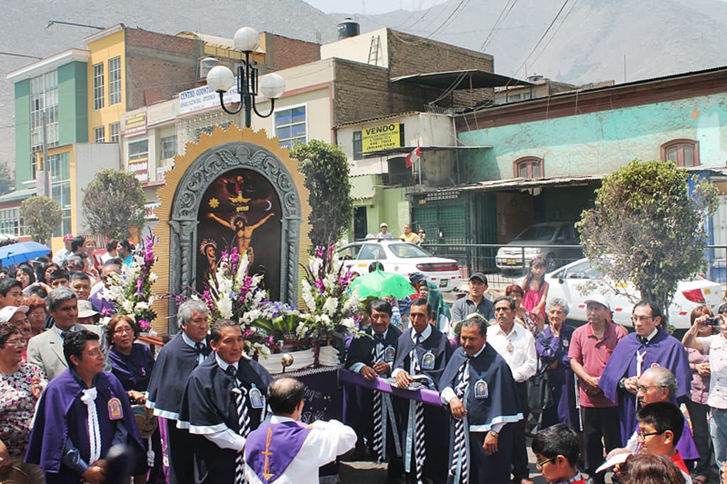 Menschen auf einer Straße mit einem Kreuz und Blumensträußen in der Mitte, Autos fahren dahinter und Gebäude, Bäume und Straßenlaternen im Hintergrund.