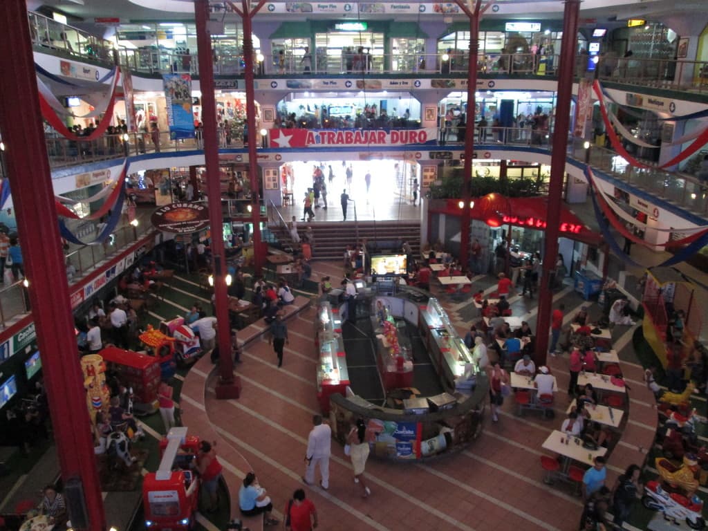 Ein belebter Einkaufszentrum-Szenen mit Säulen, Menschen, Tischen, Fliesen, Flaggen, Geschäften, einem Banner, Treppen und einem Namensschild.