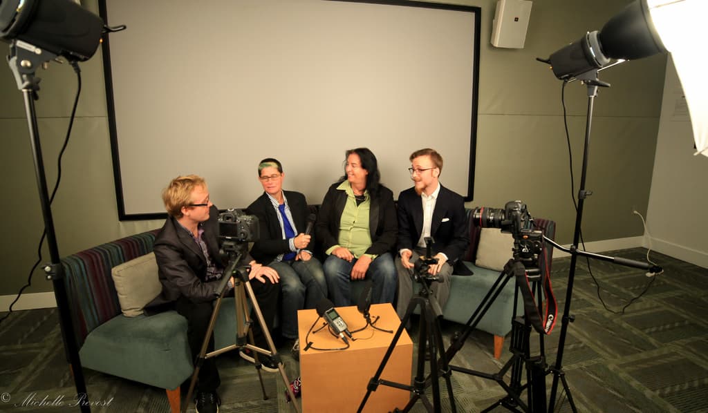 Eine Gruppe von drei Personen sitzt auf einem Sofa und gibt ein Interview, wobei eine Person in einem schwarzen Mantel und Hose links, eine Person in einem Mantel und blauer Krawatte mit einem Mikrofon in der Mitte und eine Person rechts lächelt. Ein Mikrofonrekorder befindet sich auf dem Tisch in der Mitte, Kameras sind auf der rechten und linken Seite und große Scheinwerfer sind vorhanden, mit einem Projektionsschirm im Hintergrund.