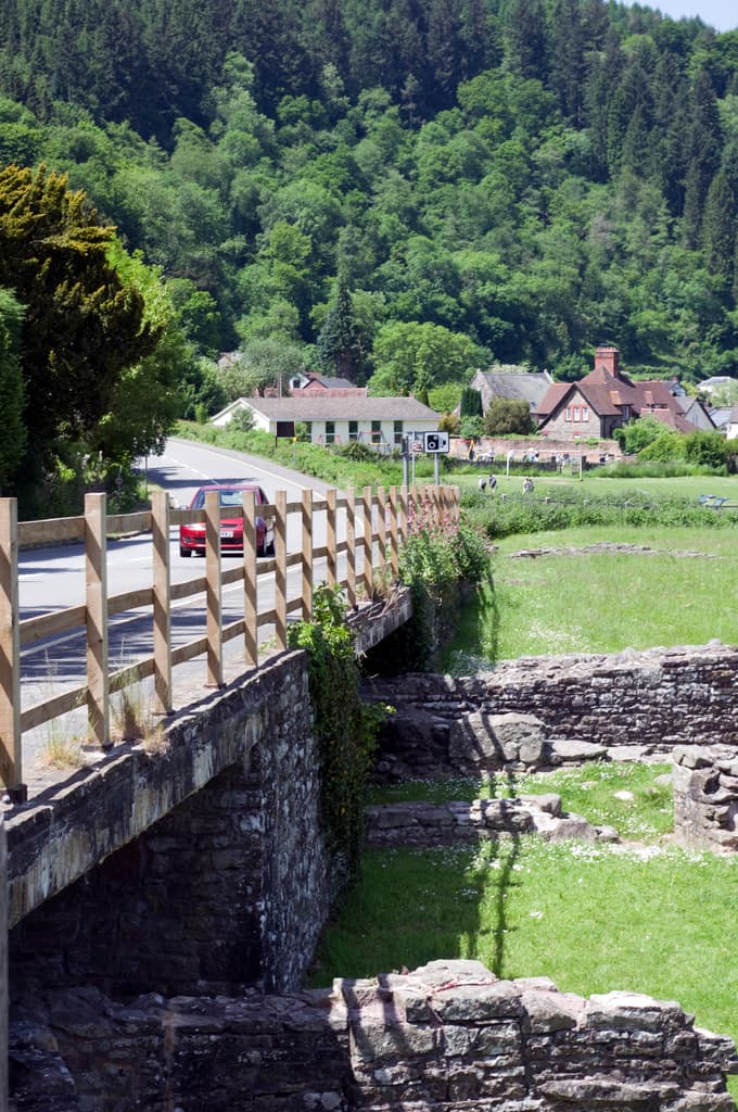 Ein Auto fährt auf einer Brücke mit Bäumen, Gras, Häusern und Steinen im Hintergrund.