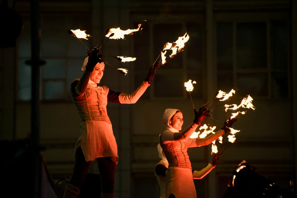 Eine Gruppe von Menschen steht in der Mitte des Bildes, jeder hält einen brennenden Feuerstab, im Hintergrund ein Gebäude.
