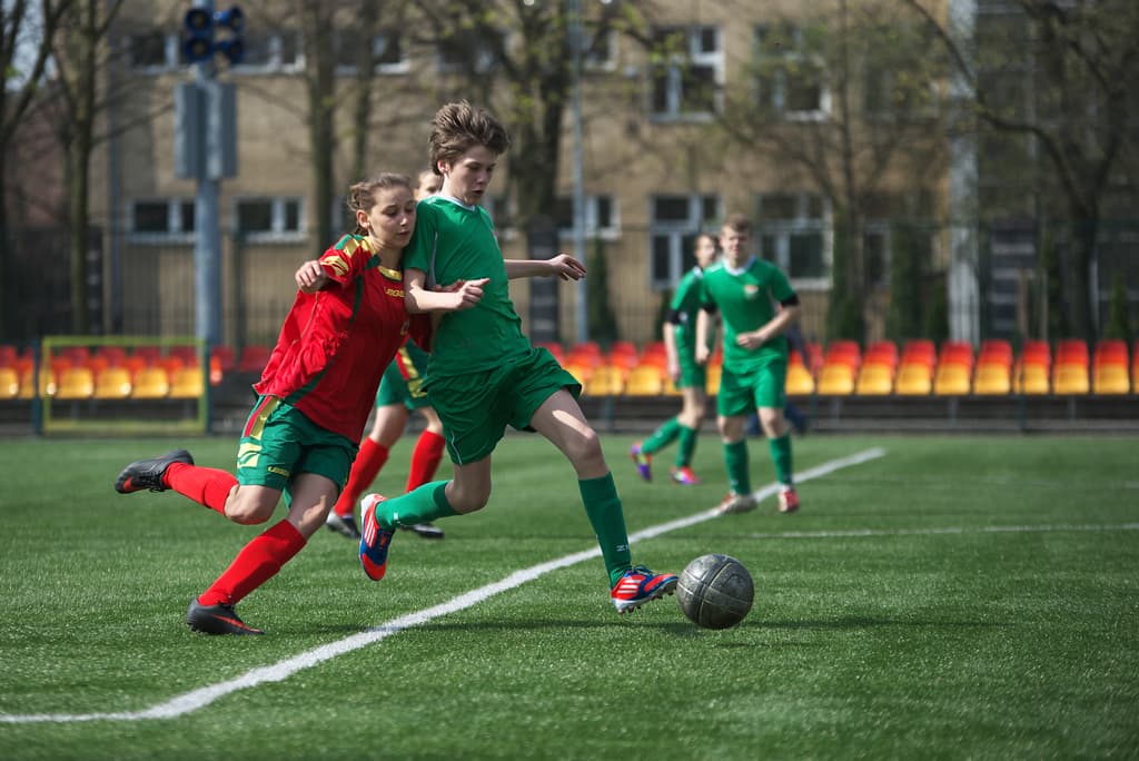 Eine Gruppe von Spielern spielt Fußball auf einem grünen Feld, umgeben von Bäumen, mit ein paar Gebäuden und Stühlen für das Publikum im Hintergrund.