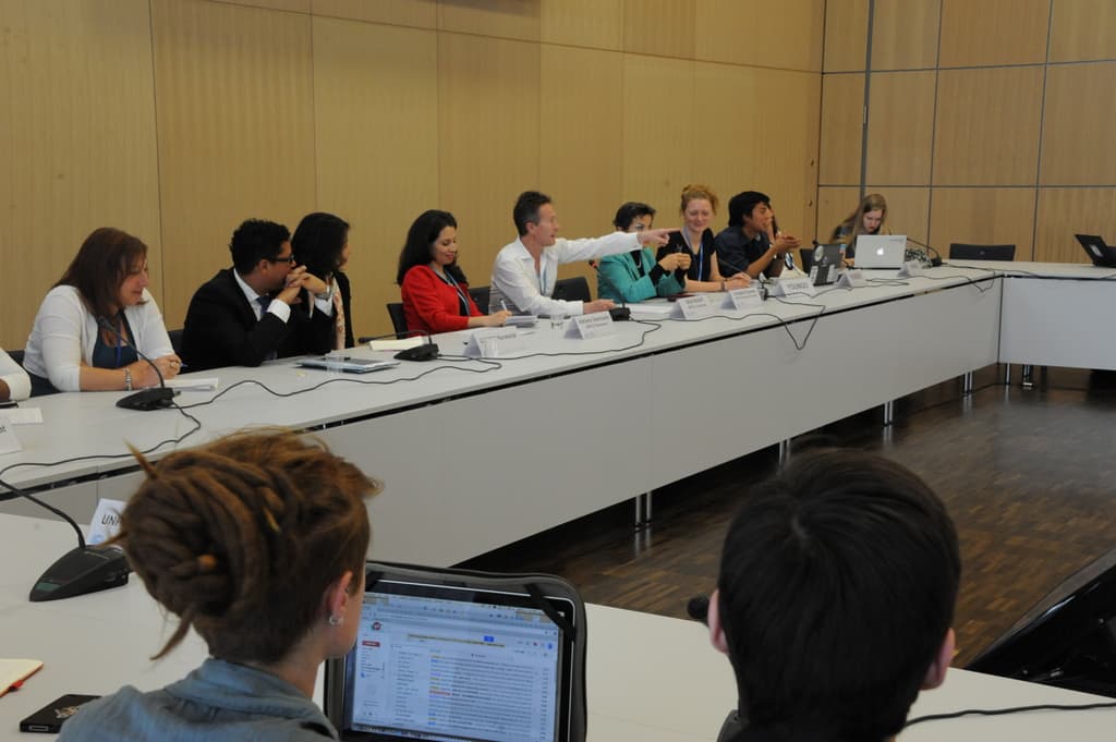 Ein Konferenzsaal mit vielen Menschen, die vor einem weißen Tisch mit Laptops, drahtlosen Mikrofonen und Namensschildern sitzen, vor einer Wand.