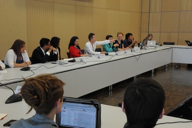 Ein Konferenzsaal mit vielen Menschen, die vor einem weißen Tisch mit Laptops, drahtlosen Mikrofonen und Namensschildern sitzen, vor einer Wand.