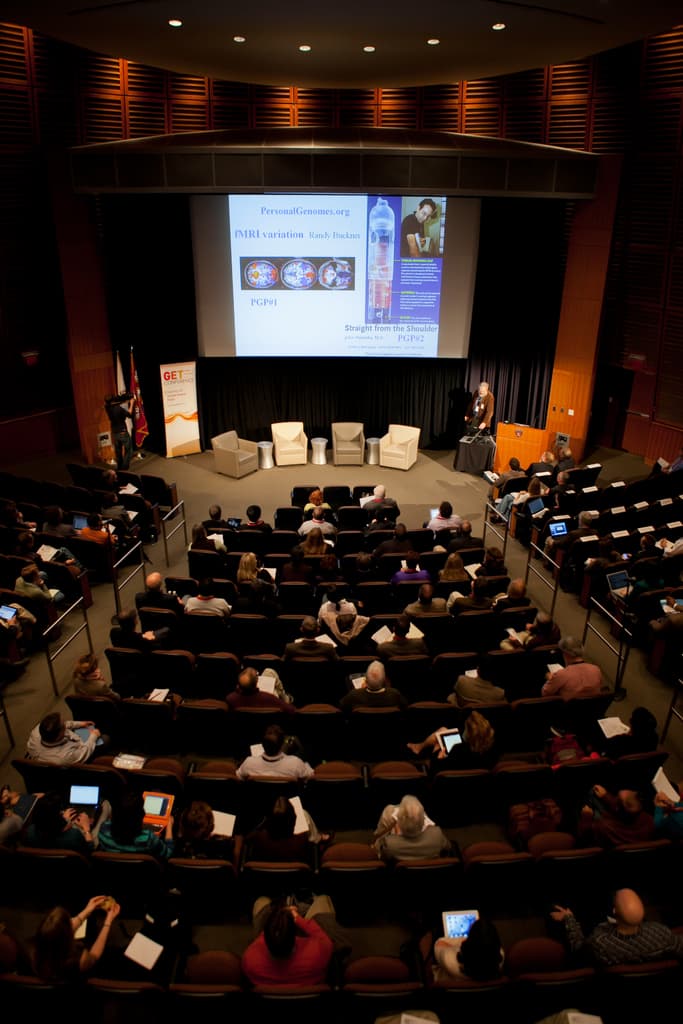 Ein mit Sitzenden und Gegenständen haltenden Menschen gefüllter Hörsaal, beleuchtet von zahlreichen Lichtern, mit Fahnen links, einem Podium rechts und einem Werbeplakat links.